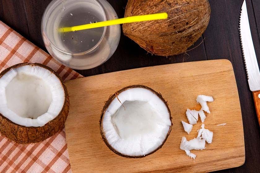 A board on which there is a coconut cut in 2, another whole one and a container of coconut juice and a straw