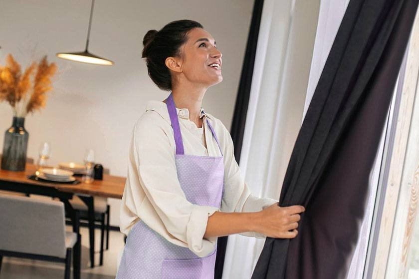 A woman in a purple apron adjusts a curtain