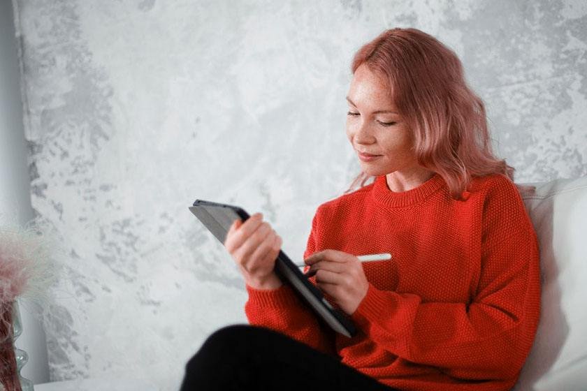 A woman in a red blouse is sitting on a couch and typing on her tablet