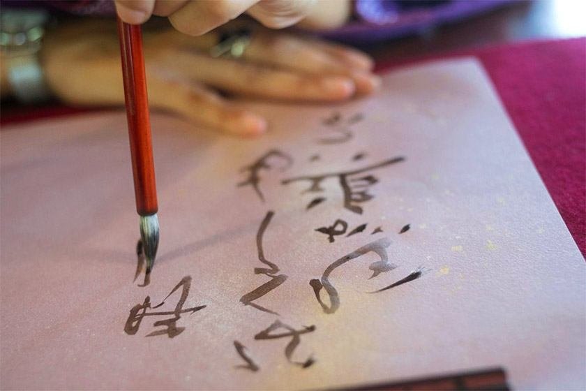 A hand holding a traditional Japanese pen writes on a piece of paper