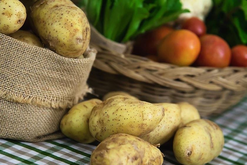 Fresh peeled potatoes on table, more potatoes in canvas bag and tomatoes and onions in background