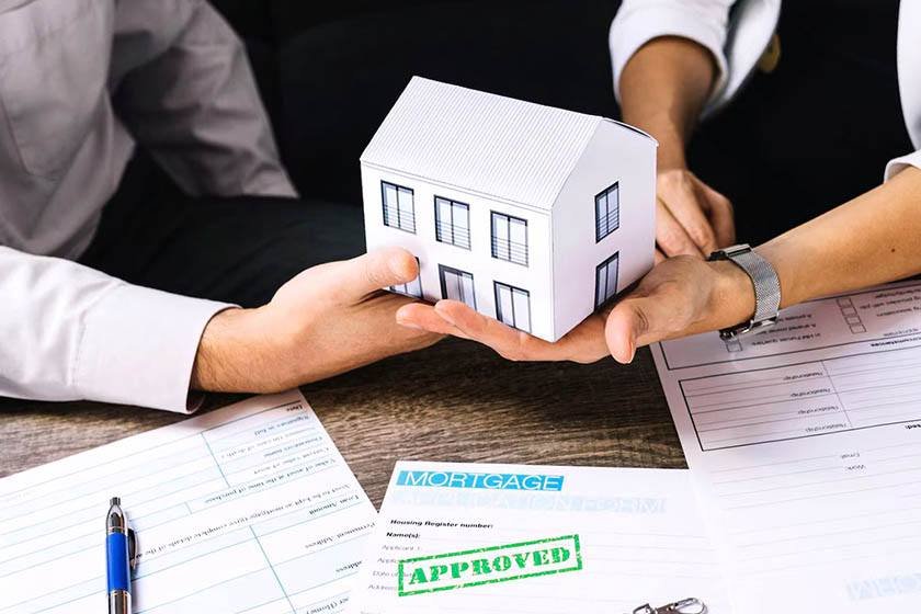 Two men hand each other a model of a house, while in front of them is a sheet of contract and a pen